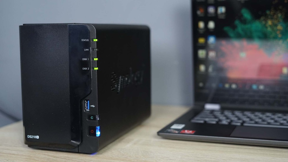 Close-up of Synology NAS with glowing LED indicators and a laptop in the background.