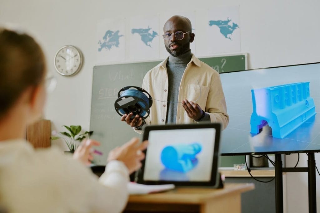 An instructor explaining 3D printing with teaching materials and a screen demonstration