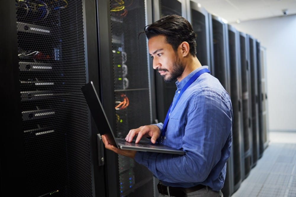 A technician working on a laptop in a server room filled with network servers, showcasing tech expertise and data management.