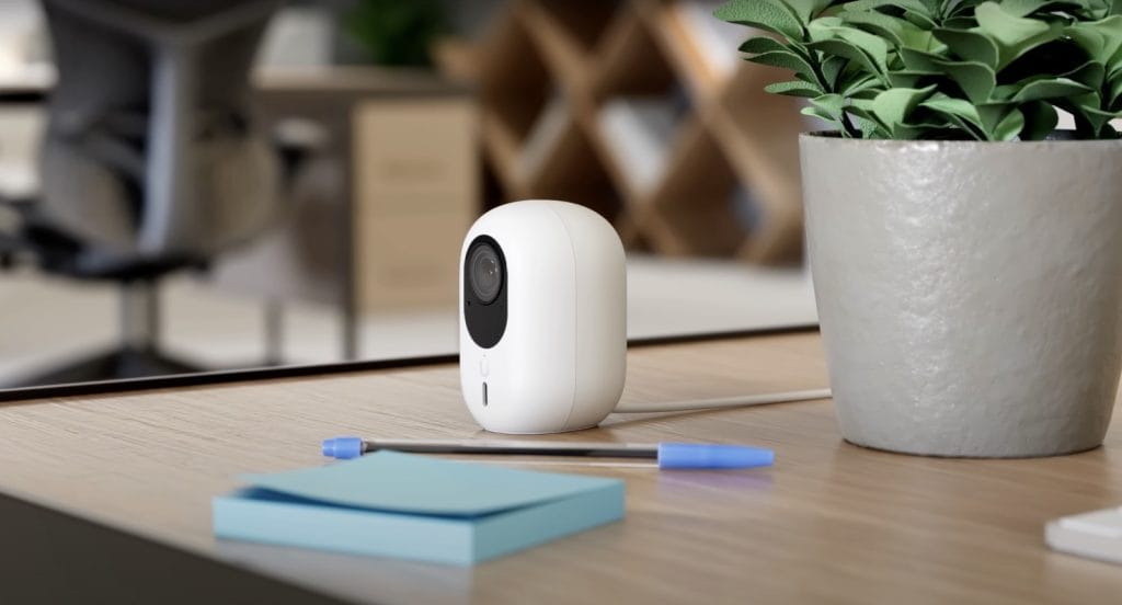 A modern white security camera placed on a wooden desk with stationery and a potted plant in the background.