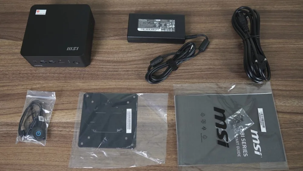 An MSI Cubi series mini PC with accessories including a power supply, cables, and a quick start guide on a wooden surface.