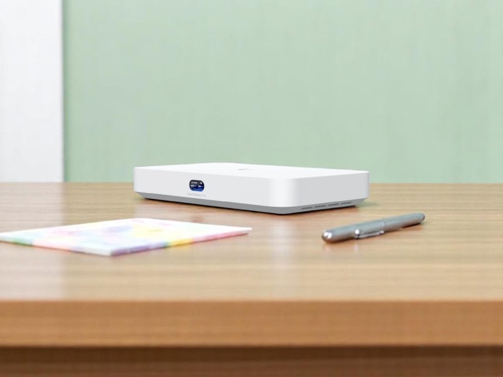UCG-Fiber. A sleek white Cloud Gateway Fiber device on a wooden table with a silver pen and a colorful notebook nearby