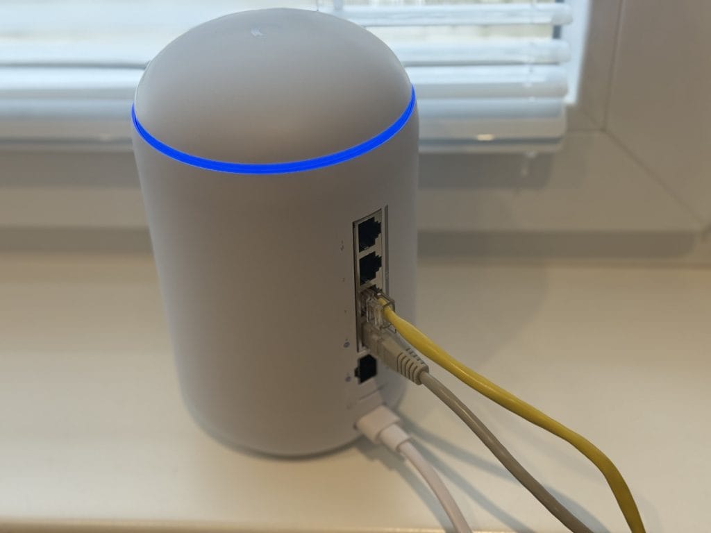UniFi Dream Router 7. Modern white cylindrical Wi-Fi router with blue LED light and multiple ethernet cables connected, placed on a white surface near a window blind