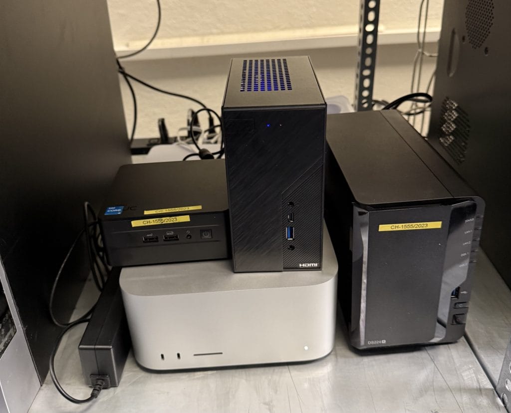 A collection of small form-factor desktop computers and a network-attached storage device stacked on a metal surface with visible cables