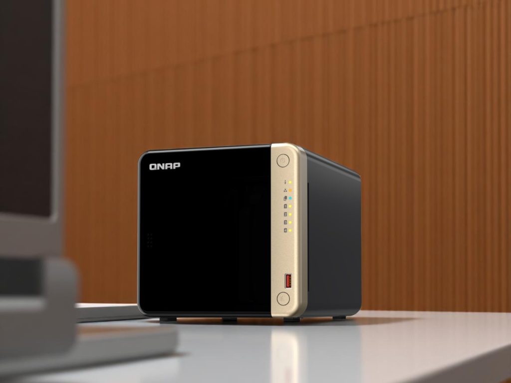 A black and gold QNAP network attached storage (NAS) device on a white desk with wooden panel background
